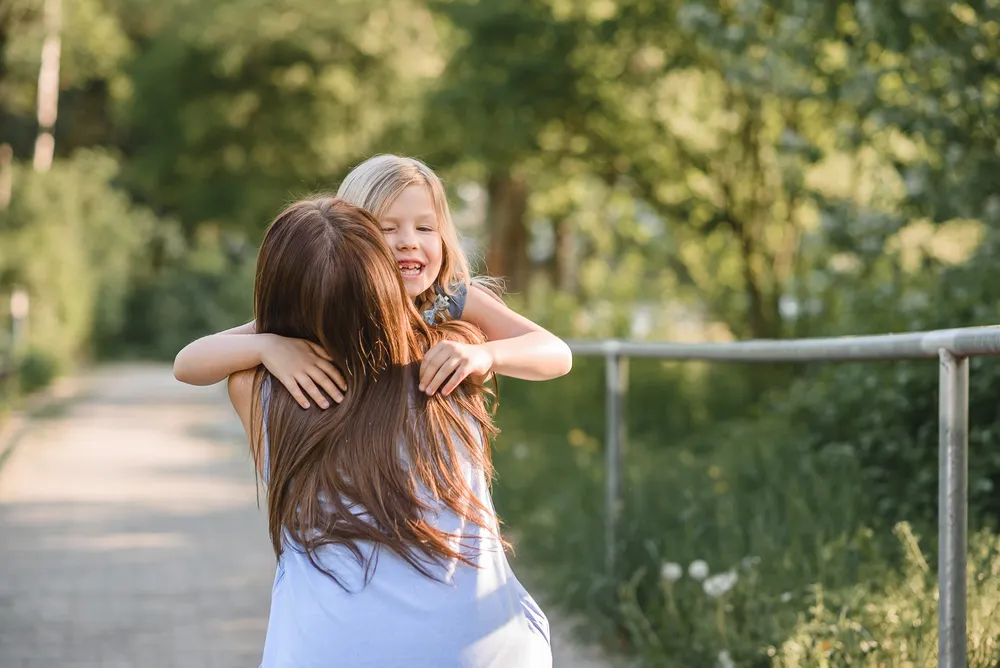 Two young girls share a joyful embrace on a sunlit park path, surrounded by greenery. The girl with blonde hair beams, while the brunette's back is turned. A metal fence lines the right side of the scene.