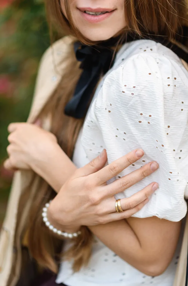 Woman in white blouse with black bow, holding sleeve, gold ring, pearl bracelet, outdoor, soft focus background.