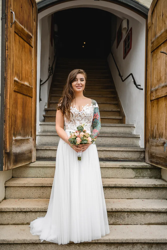 Woman in white wedding dress stands on stone steps, holding bouquet, with tattooed arm visible. Open wooden doors behind her lead to dark interior.