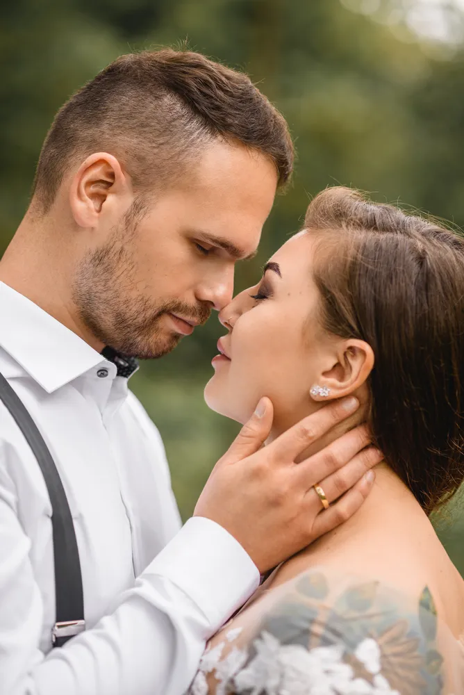 Man and woman share a tender moment outdoors. He gently cups her face, she smiles softly. He wears a white shirt with black suspenders; she dons a floral dress. Blurred greenery forms the backdrop, highlighting their intimate connection.