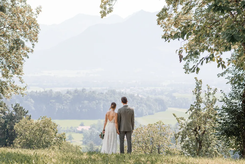 Couple in wedding attire stands on a grassy hill, gazing at a misty mountain range. Lush greenery surrounds them, with trees framing the view. Serene and romantic atmosphere.
