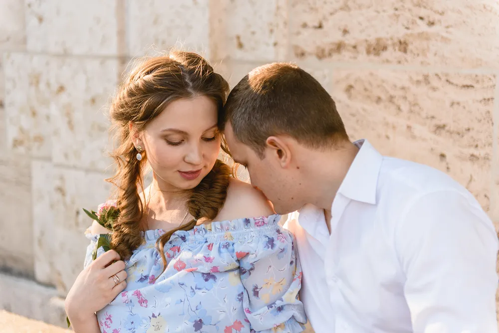 Couple sharing a tender moment, woman in floral off-shoulder dress holding a rose, man in white shirt, against a textured stone wall.