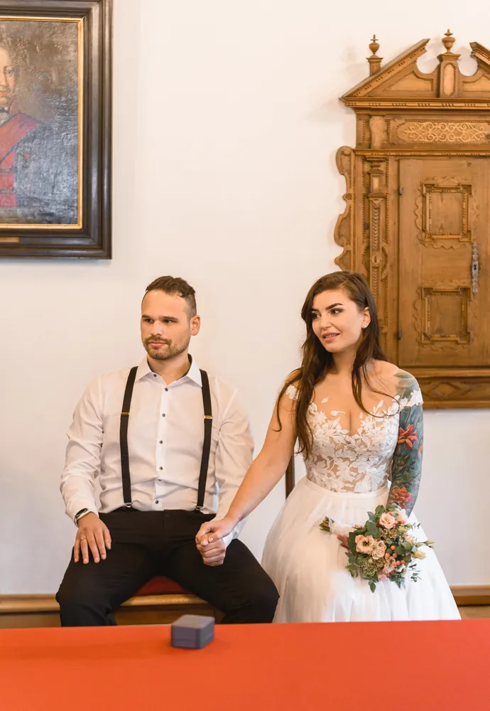 Couple seated at table, holding hands, in formal attire, woman holding bouquet, ornate wooden cabinet and painting in background.