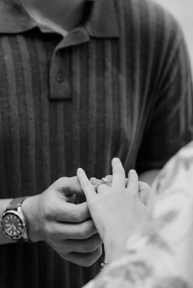 A person wearing a striped polo shirt receives a ring on their left hand from another individual, with a watch visible on the left wrist. The background is indistinct, focusing the viewer's attention on the exchange.