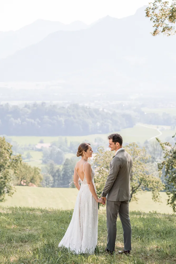 Bride in white gown and groom in gray suit hold hands, smiling, on grassy hill overlooking misty valley with trees and distant mountains.