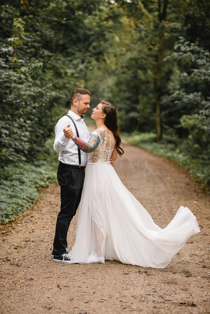 Couple in wedding attire dancing on a dirt path surrounded by trees. Woman's dress flows, man wears suspenders and sneakers. Romantic, natural setting.