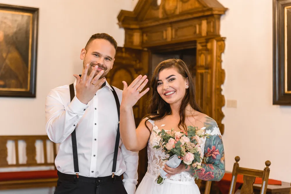 Bride and groom in white attire, both wearing wedding rings, smiling and holding up hands to display rings. She holds a bouquet of pink and white flowers. They stand in a room with wooden furniture and classical decor. No other people are visible.