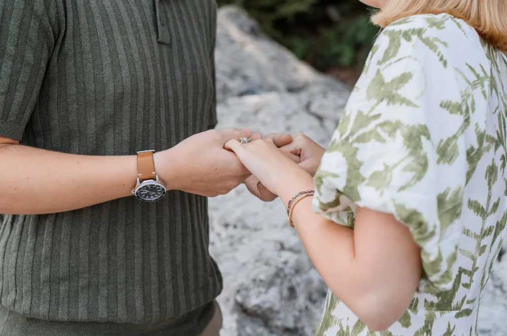 A couple shares an intimate moment outdoors, with the man presenting a ring to the woman. He is dressed in a green striped polo and wears a brown leather watch, while she is adorned in a white blouse with green leaf patterns. They stand against a backdrop of a rocky landscape, with the woman's blonde hair catching the light. The focus is on their hands, where the ring is being placed on her finger, signifying a commitment.