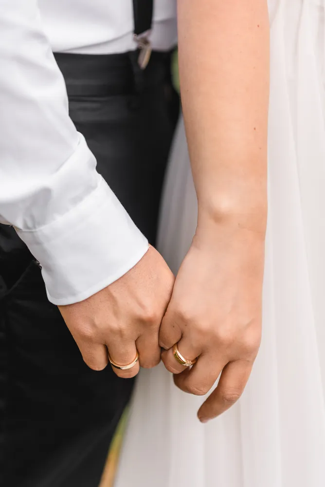 Couple's hands clasped, wedding rings visible. Man in white shirt with black trousers, woman in white dress. Outdoor setting.