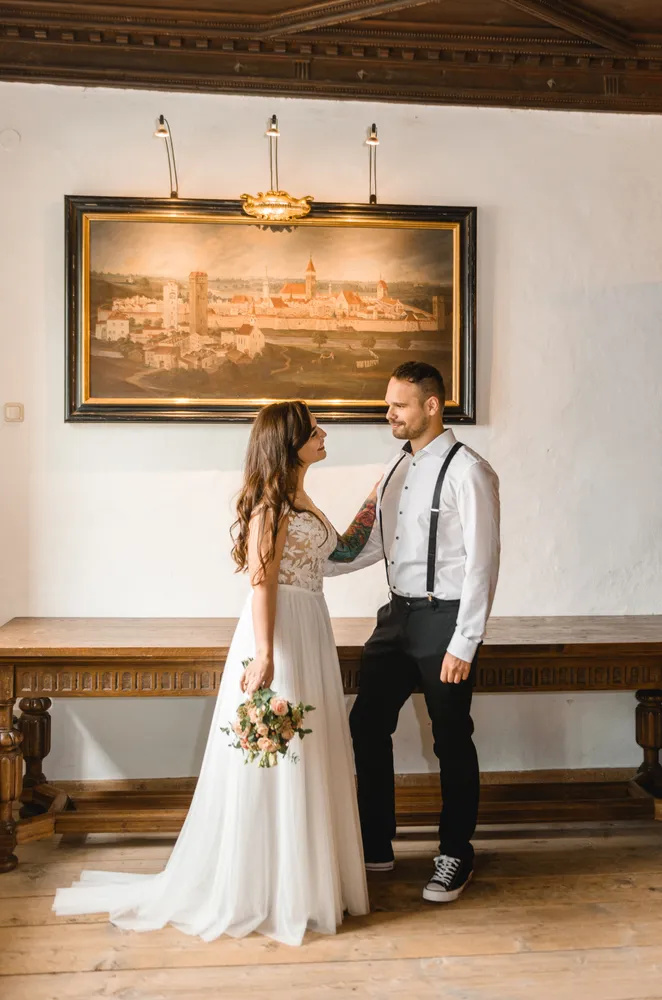 Bride and groom embrace in a historic hall. She wears a white lace gown, holding a floral bouquet. He is in a white shirt, black suspenders, and casual sneakers. A large framed cityscape painting adorns the wall behind them.