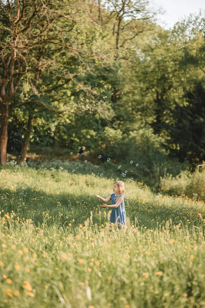 Girl in blue dress blows soap bubbles in sunlit meadow surrounded by trees.
