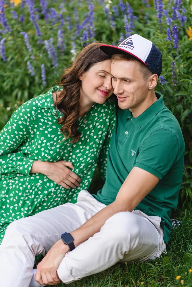 Couple sitting closely in a grassy area with blooming purple flowers. Woman in green floral dress leans on man's shoulder. Man wears green polo, white pants, and a white cap. Both smiling softly, woman's hand on her stomach. Peaceful, affectionate pose.