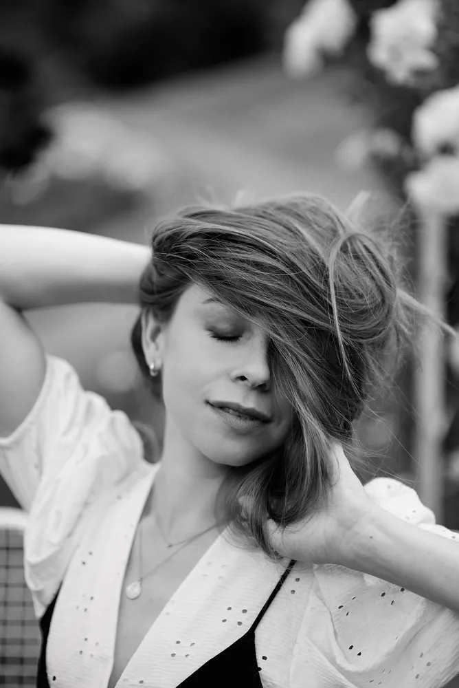 Woman with closed eyes and a serene expression, hair gently tousled, wearing a white blouse with a black garment underneath. Outdoors, with soft focus on foliage in the background.