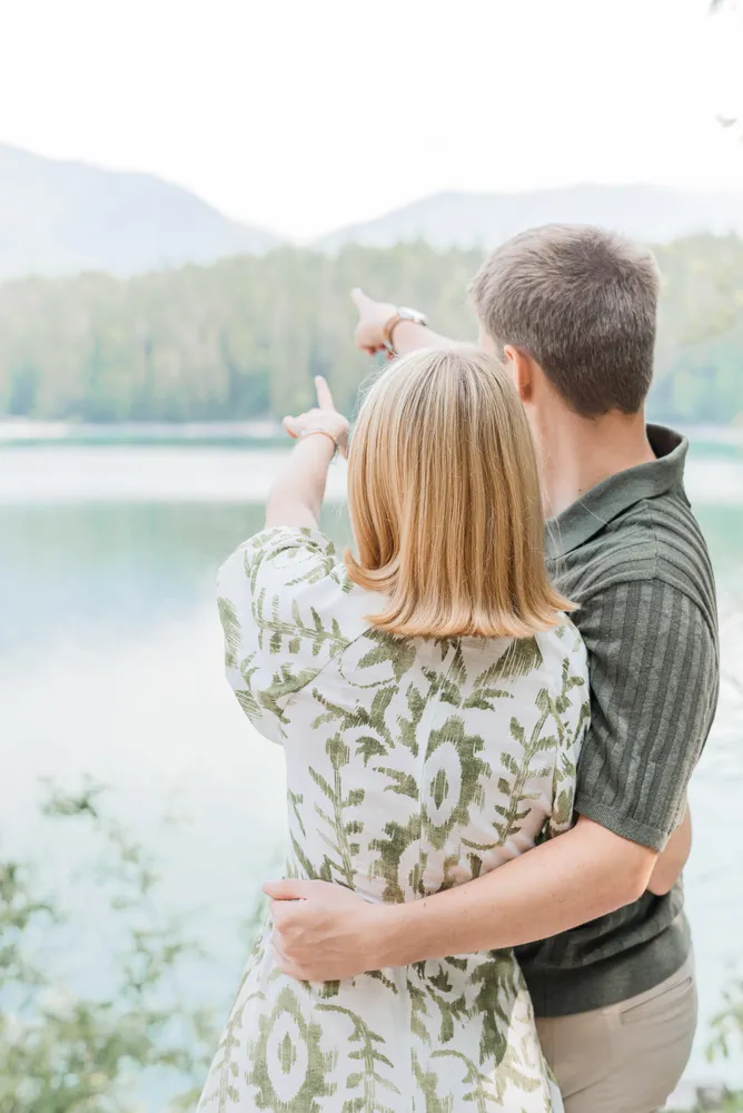 Couple points towards scenic lake, embracing in natural setting. Woman in green-and-white patterned dress, man in dark-striped polo. Trees and mountains visible in background. No faces shown.