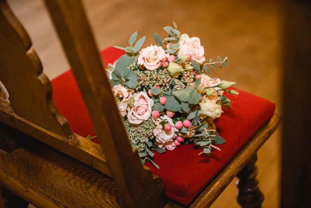 A bouquet of pink roses, white flowers, and green foliage rests on a red velvet cushion atop a wooden chair, set against a blurred wooden floor background.