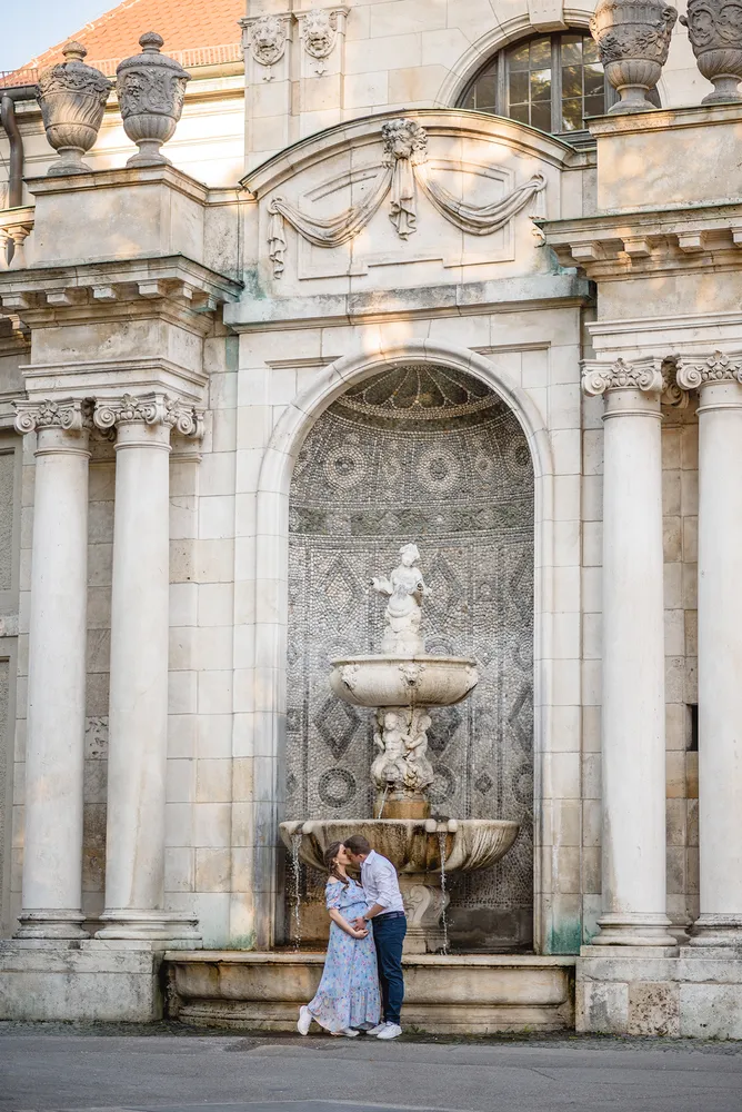 Couple shares a kiss in front of an ornate stone fountain with classical architecture, under soft daylight.