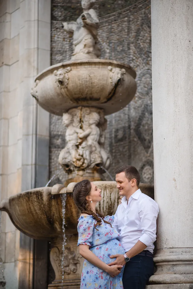 Expectant couple smiles at each other before a grand fountain adorned with sculptures, set against an ornate building façade.