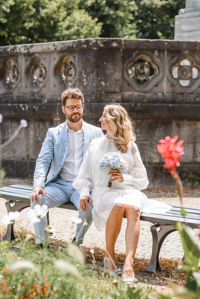 Couple in light blue suit and white dress sit on bench, holding bouquet, surrounded by garden and stone wall.