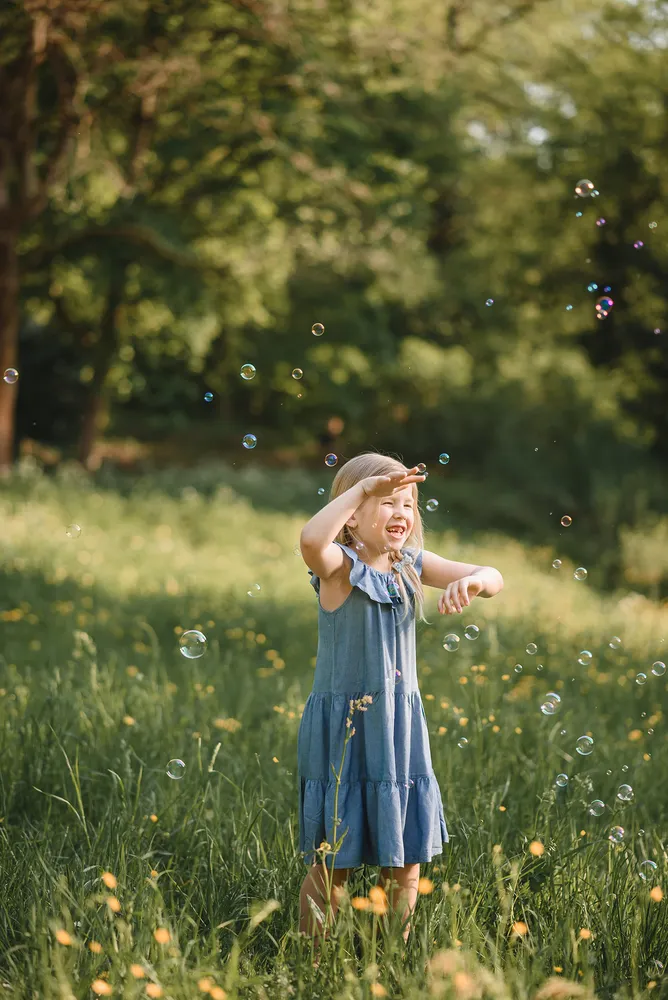 Girl in blue dress laughs while reaching for soap bubbles in sunlit meadow surrounded by greenery.