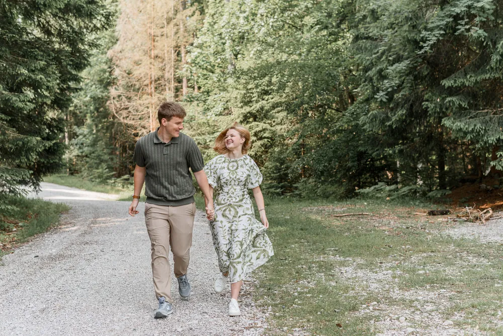 A man and woman stroll hand-in-hand along a gravel path in a sun-dappled forest. He wears a striped polo and khaki pants; she is in a green floral dress. Both are smiling, engaged in conversation.