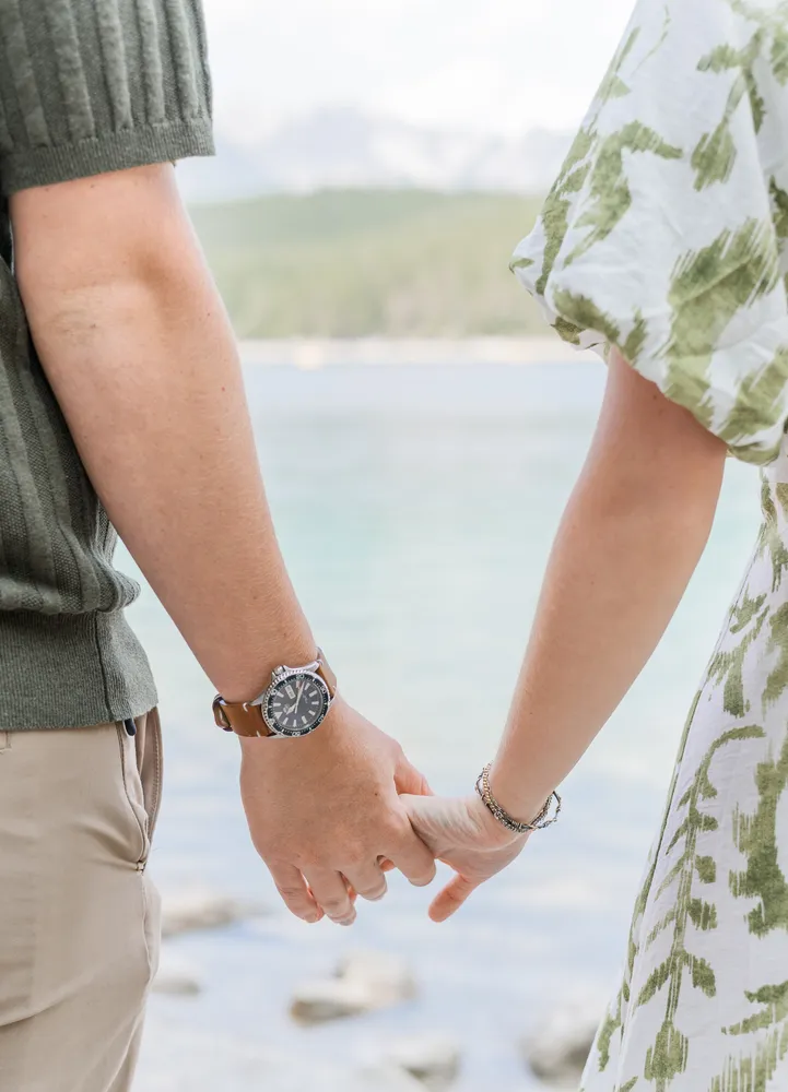 Couple holding hands by water, man in green sweater and khaki pants, woman in white dress with green pattern, man's watch visible, serene natural setting.
