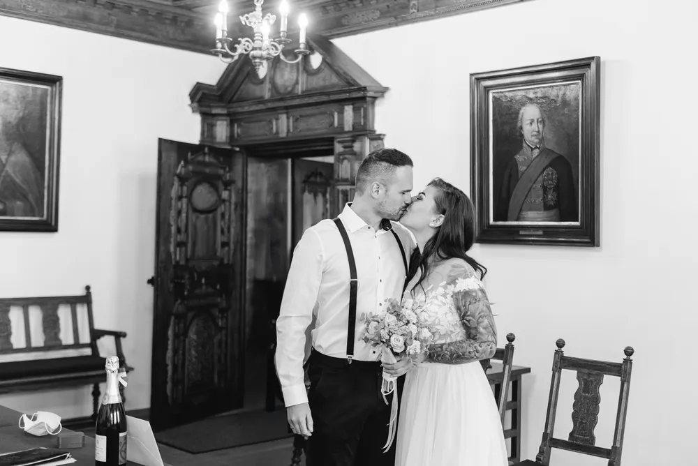 Couple shares a kiss; bride holds bouquet. They stand in a room with vintage decor, including a chandelier and framed portraits.