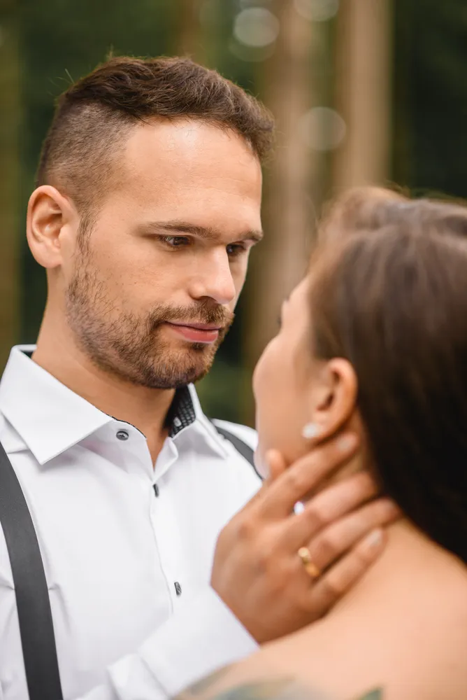 A man and woman embrace outdoors. He wears a white shirt with black suspenders, she has brown hair. He gently holds her face, both smiling softly.