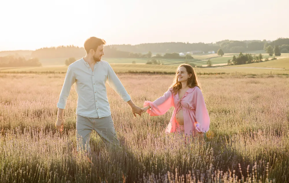 Expectant couple walks hand-in-hand through a field of purple flowers at sunset. Man in white shirt, woman in pink dress showing baby bump. Serene rural landscape in background.