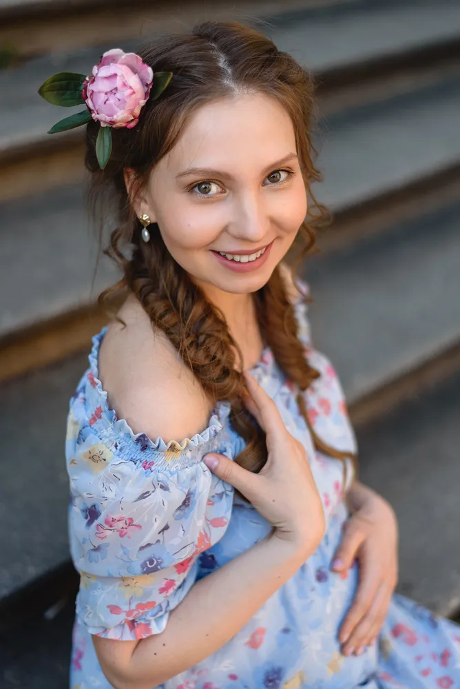 Woman in floral dress smiles, hand on shoulder, adorned with pink flower in hair, seated on stone steps outdoors.
