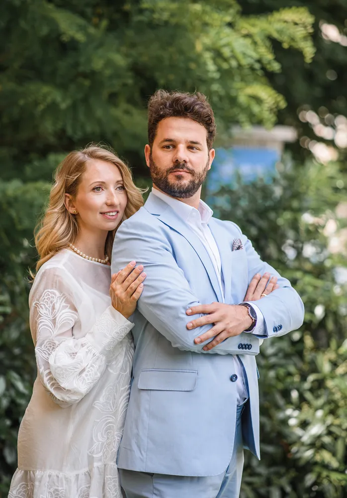 Couple in formal attire stands outdoors, woman in lace dress with pearls, man in light blue suit, arms crossed, amidst greenery.