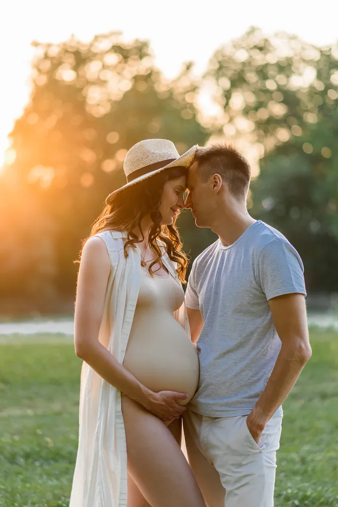 Expectant couple in serene embrace; pregnant woman in white robe and hat, man in grey shirt and khaki pants, tenderly touching foreheads in sunlit park.
