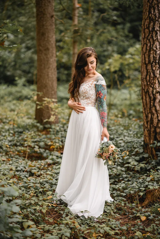 Woman in white lace gown stands in forest, holding floral bouquet, tattooed arm visible.