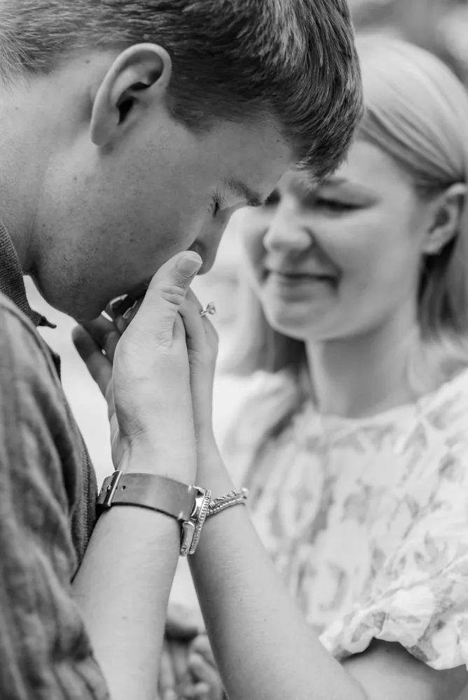 A man receives a ring from a woman in a black-and-white photograph. He looks down at her hand, which holds the ring close to his face, while she smiles softly. The setting is blurred, focusing on their intimate moment.