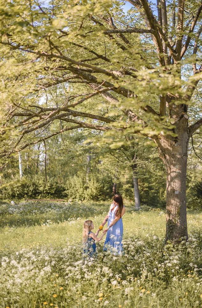 Two women in blue dresses share a sunflower under a blooming tree in a verdant meadow.