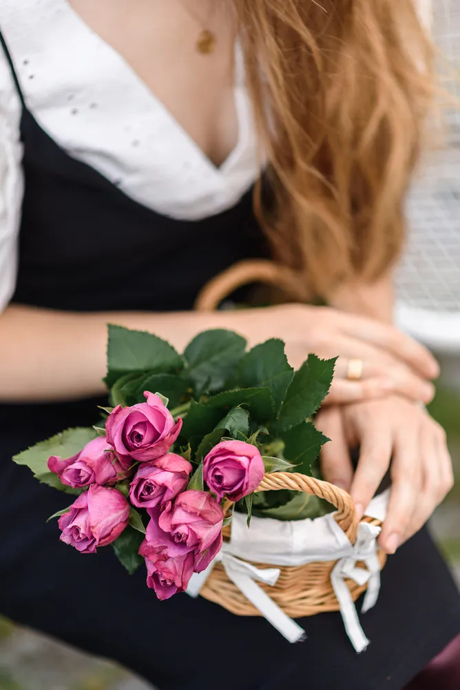 Woman in white blouse and black dress holds pink roses in wicker basket, her hands adorned with a ring, outdoors.