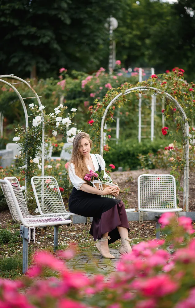 Woman in white blouse and black skirt sits on white bench, holding bouquet of pink and white flowers, surrounded by rose arches in garden, cobblestone path, blurred pink flowers in foreground.