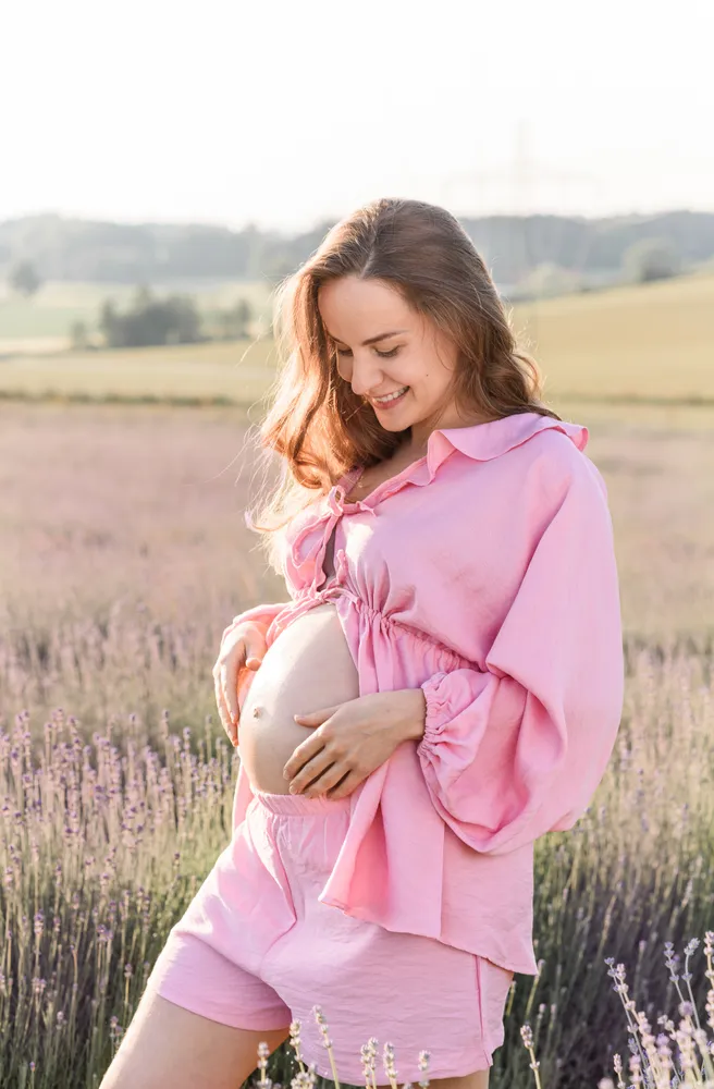 Expectant woman in pink, gently cradling her belly amidst a lavender field at sunset.