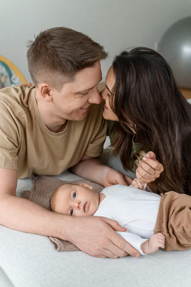 A man and a woman in casual attire affectionately interact with a baby on a light-colored bedspread. The man's arm supports the baby's back while the woman's hand gently touches the infant's hand. The baby, dressed in a white long-sleeve onesie and brown pants, gazes upwards. The couple leans in to kiss the baby's forehead, conveying a sense of familial bonding. The setting appears to be a domestic interior with neutral tones.