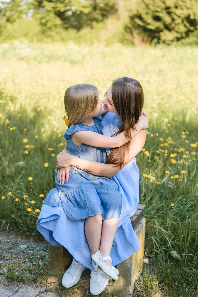 Mother and daughter in blue dresses embrace on a bench, surrounded by a sunlit field of yellow flowers, with trees in the distance.