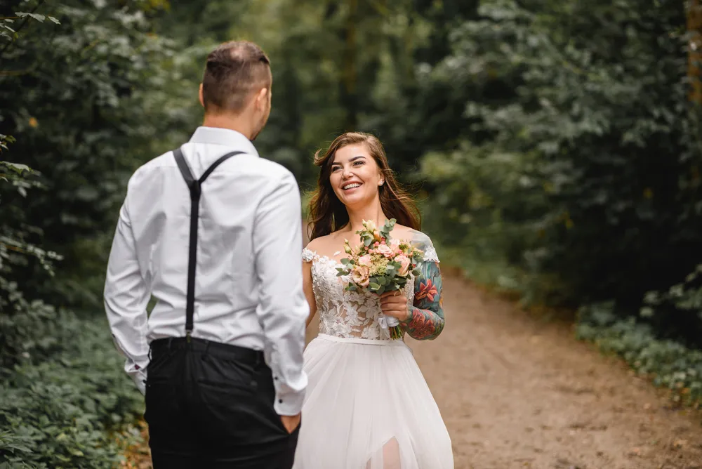 Bride and groom embrace in a forest clearing, she holds a bouquet, he wears suspenders, both dressed formally, natural light, joyful expressions.