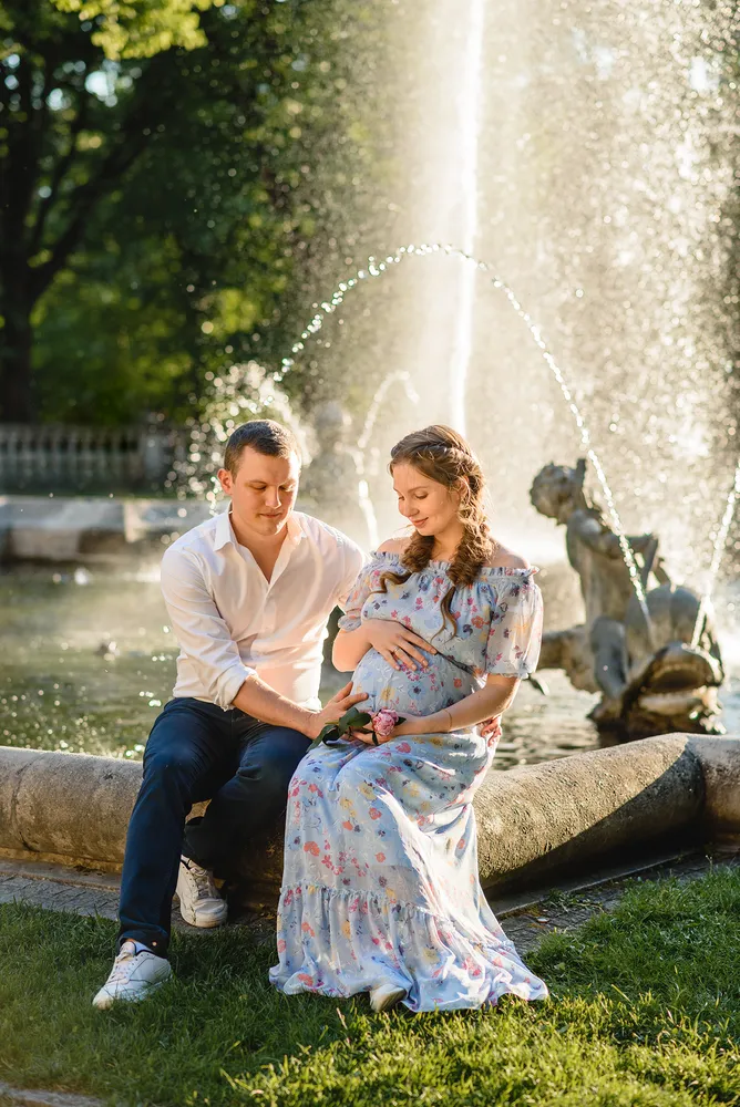Couple sits by fountain; woman in floral dress cradles belly, man in white shirt supports her. Sunlit park, statue and greenery in background.