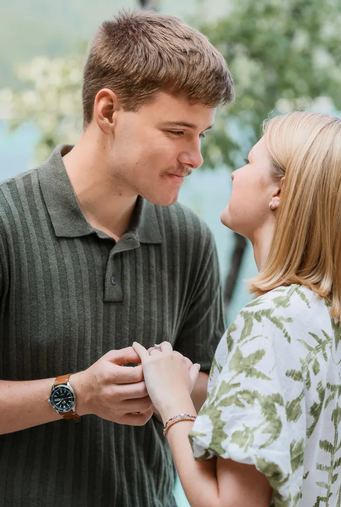 A young couple smiles while holding hands outdoors. The man wears a striped polo and watch; the woman is in a green floral top. Blurred greenery suggests a park setting.