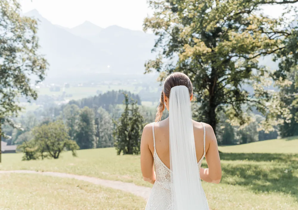 Bride in lace wedding dress stands on a grassy hill, facing a scenic valley with mountains in the distance. A dirt path winds through the green landscape.