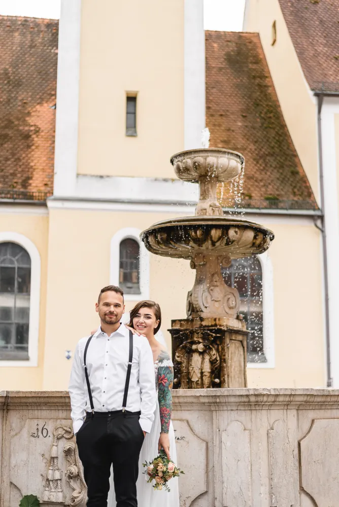 A couple in wedding attire stands before an ornate stone fountain, set against a historic building with a mossy roof. The man wears a white shirt, black pants, and suspenders. The woman is in a white dress with floral sleeves, holding a bouquet. They are smiling and gazing at the camera.