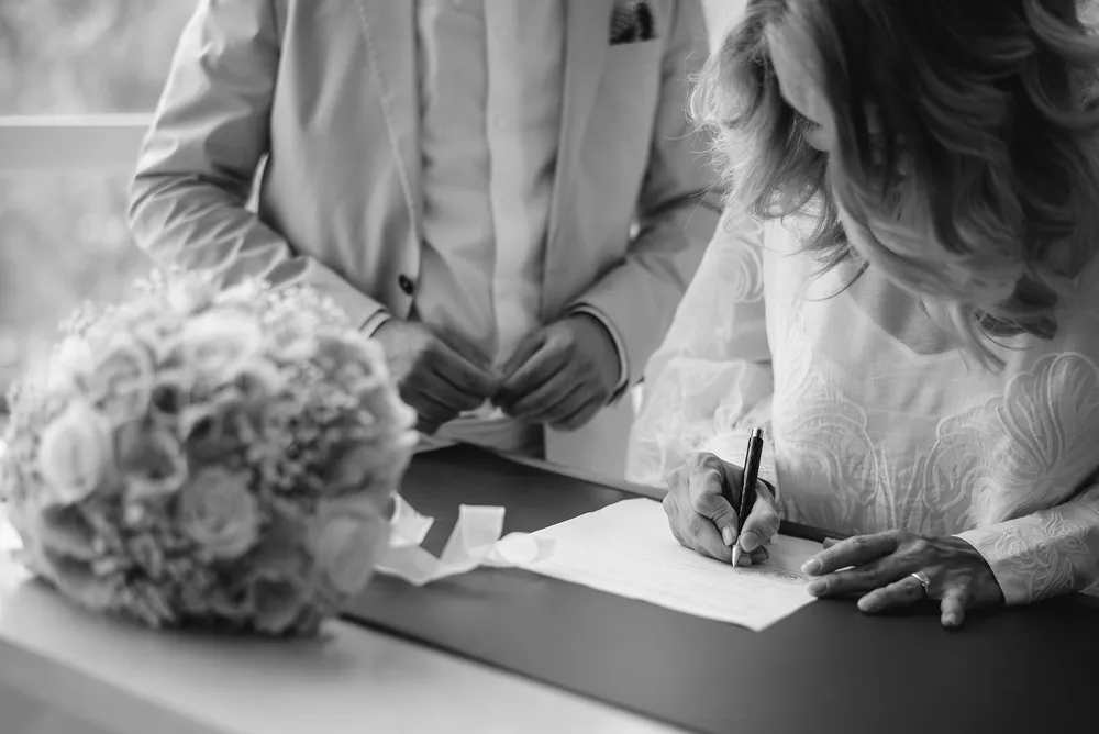 Elegantly dressed couple signs documents at a table with floral arrangement in black and white photograph.