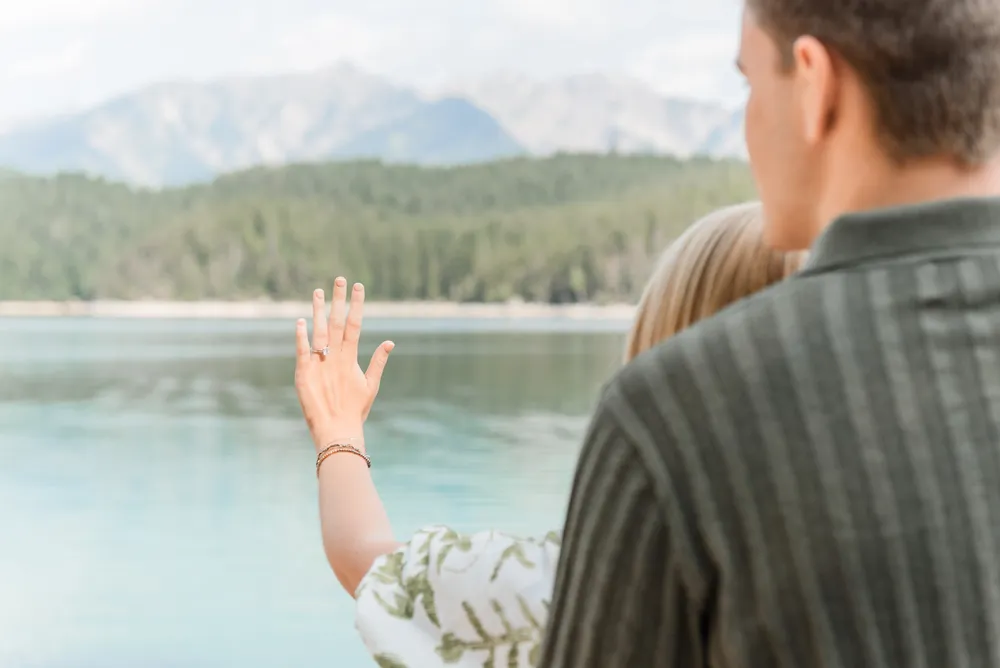 A woman with a ring on her left hand extends her arm towards a tranquil lake, while a man in a striped shirt stands beside her. They are surrounded by a serene natural landscape with mountains in the distance.