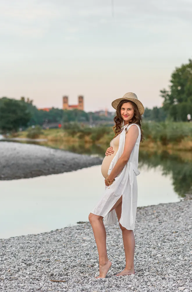 Pregnant woman in white dress and hat poses by river, city skyline in background.