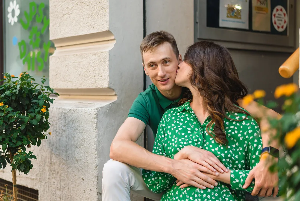 A couple shares an affectionate moment on a city sidewalk. The man, in a green shirt, kisses the woman's cheek, who wears a matching green blouse. They are surrounded by urban elements, including a textured wall and a potted plant with yellow flowers. The scene captures a candid display of intimacy in a public space.