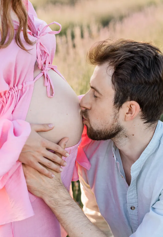 Expectant couple outdoors, man kissing pregnant woman's belly, both in soft-focus pink and blue attire amidst blurred natural backdrop.