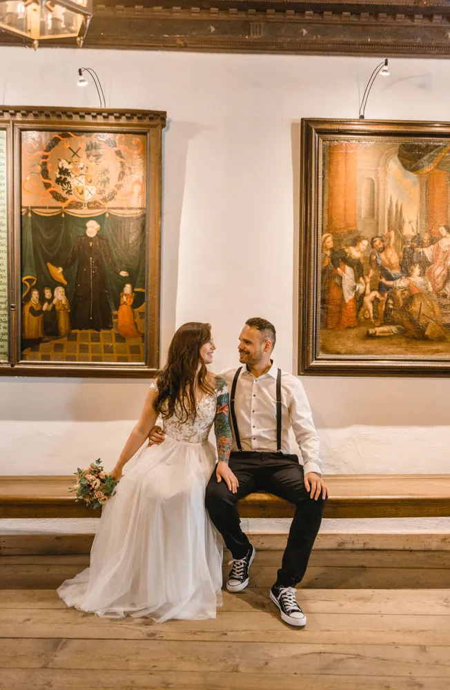 Bride and groom seated on a wooden bench in a church, bride in lace gown with bouquet, groom in white shirt and suspenders. Behind them, two religious paintings adorn the white wall. Both smiling at each other, creating a joyful wedding scene.